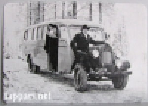 Harmaasävyinen, vanha valokuva talvitiellä seisovasta bussista, jonka luona poseeraa mies ja ovensuussa nainen.