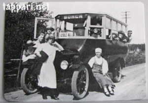 Harmaasävyinen, vanha valokuva bussista, sen edustalla seisovista naisista, ja maantiestä.